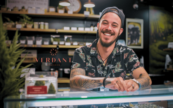 Smiling cannabis dispensary employee with tattoos and floral shirt standing behind a glass counter, with cannabis products displayed in the background and the Verdant Strategies logo overlaid.