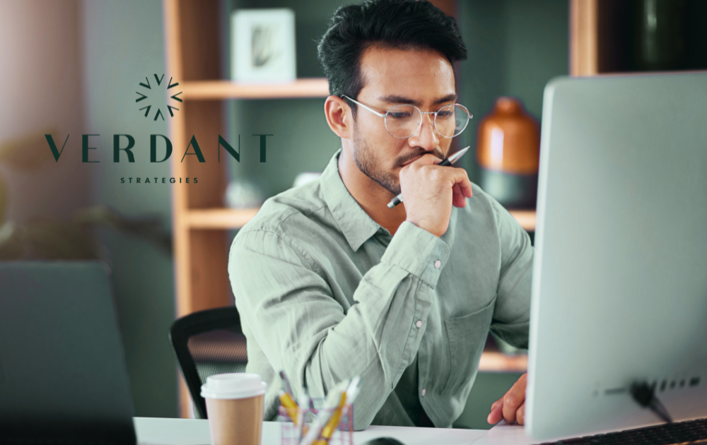 Professional man wearing glasses and a green shirt concentrates while working on a desktop computer in a modern office setting. A pen rests against his lips, and a coffee cup and office supplies sit on the desk. The Verdant Strategies logo appears on the left side of the image.