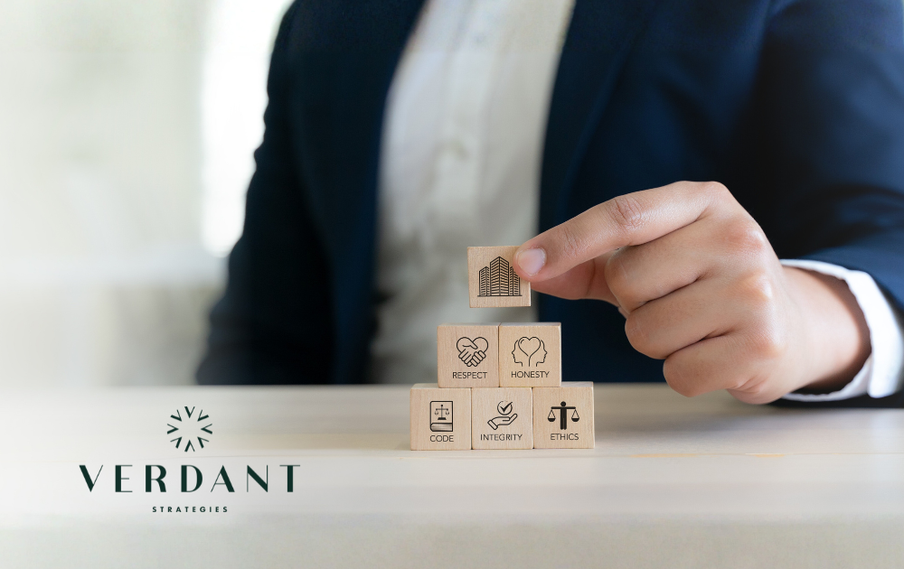 Person stacking wooden blocks symbolizing ethics, integrity, respect, and honesty, with Verdant Strategies logo in corner.